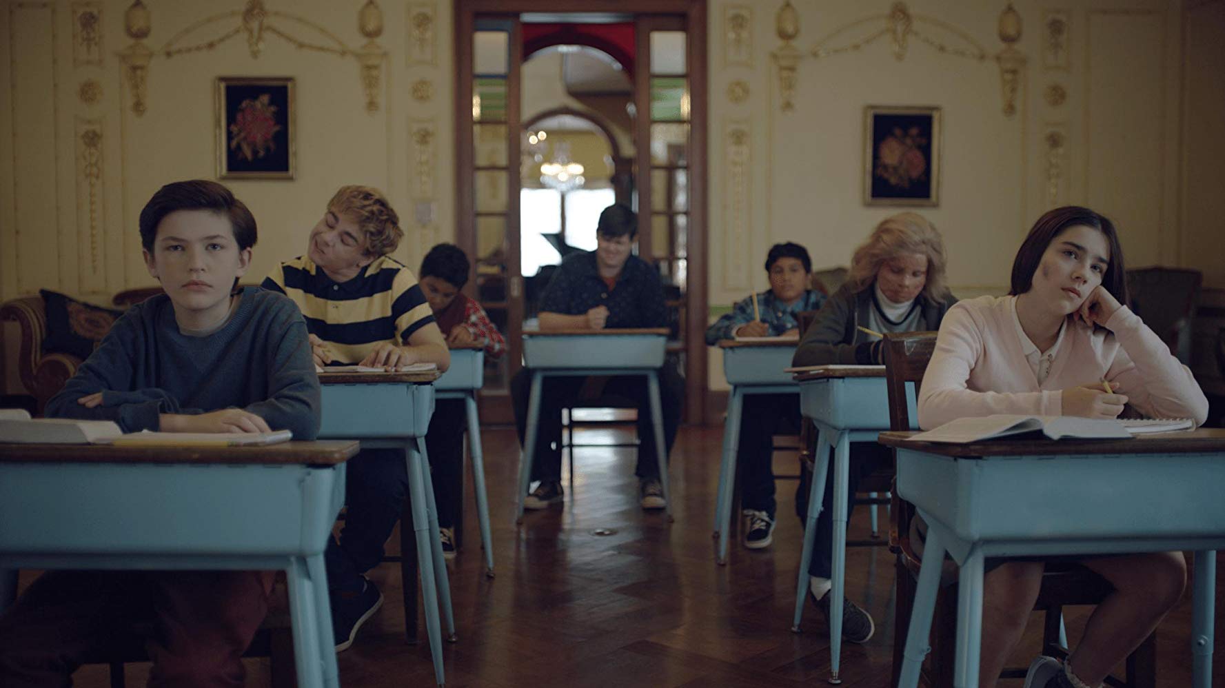 Students sitting in desk-chair combos looking forward and slightly bored. To the left is a boy with pale skin and long-ish brown hair (Jacob), to the right is a pale girl with long brown hair with her chin resting on her fist (Isabel). The desks are blue and the walls are yellow wallpaper with a Victorian-looking pattern on them.
