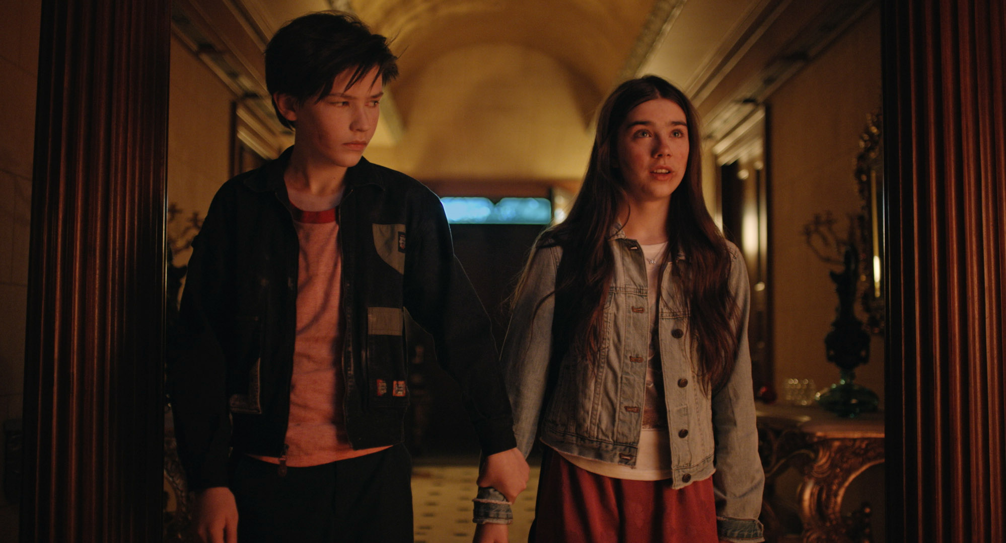Jacob and Isabel stand together in a hall lit with red-orange light. He is looking at her and holding onto her wrist, while she looks ahead with a shocked/scared expression.
