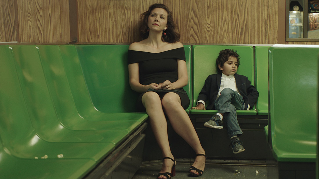 Maggie Gyllenhaal, a thin white woman with short dark hair and wearing a black dress, is looking into the distance to the left. A small child sitting next to her with his legs crossed, looking in the opposite direction. The chairs are green plastic and the walls are a medium-tone wood with prominent grain.