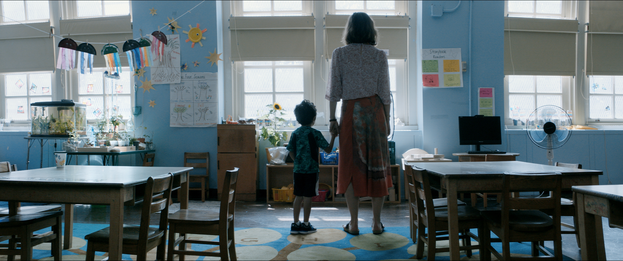 A white woman in a long skirt and white blouse stands with her back to the audience, holding hands with a small child with dark hair, both looking out a window in what appears to be a classroom.
