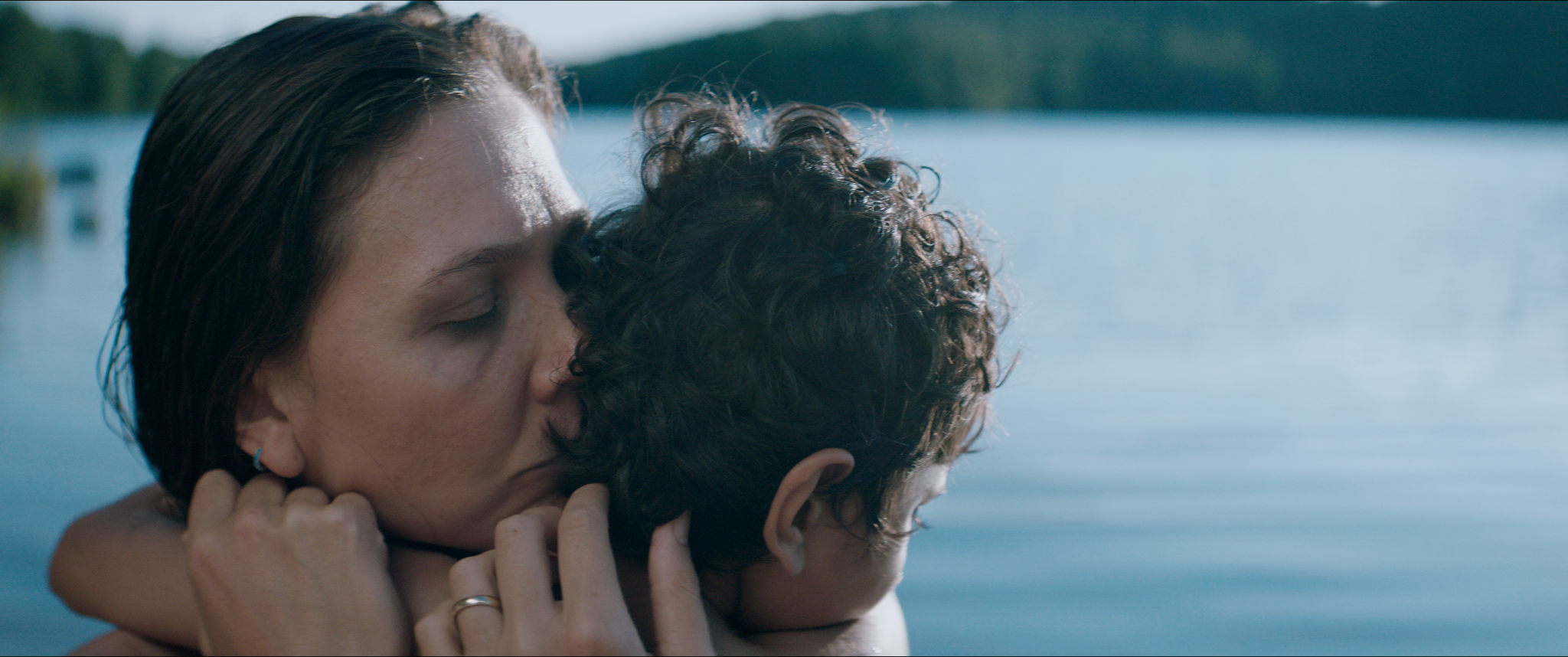 Maggie Gyllenhaal holds Parker Sevak in a tight embrace, her face turned towards his hair. There is a lake in the background.
