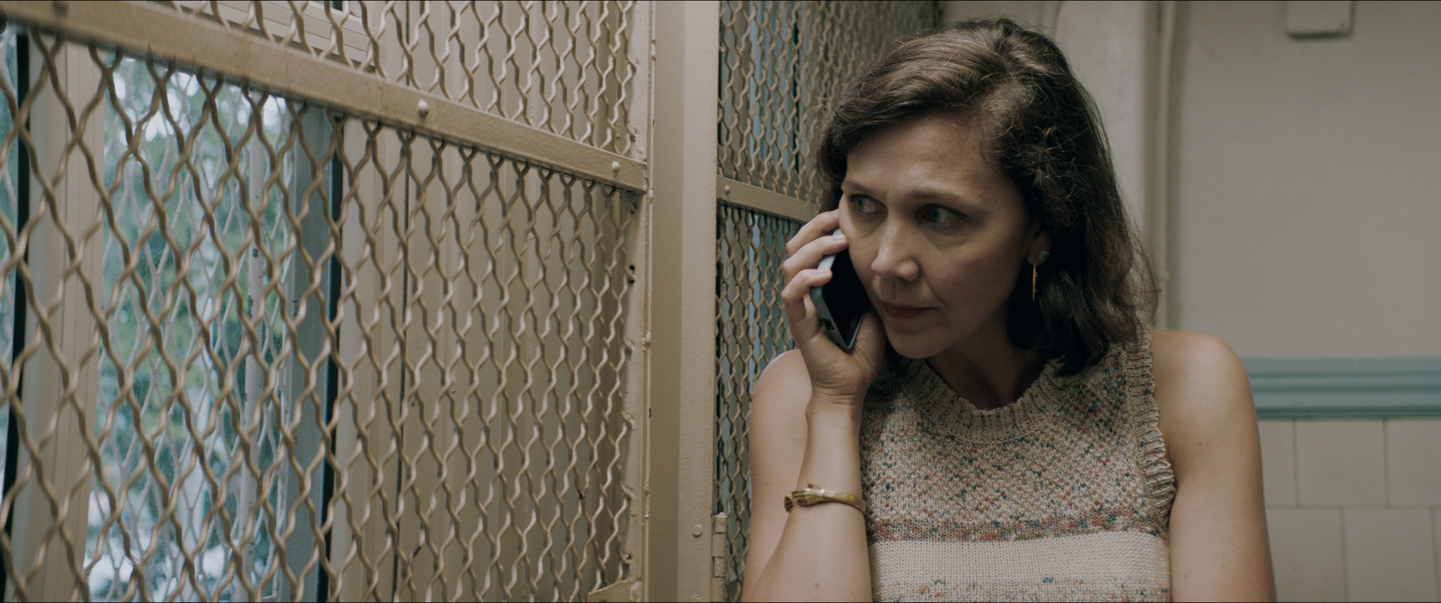 Maggie Gyllenhaal, wearing a beige sleeveless sweater, is holding a cell phone, looking to her left through a beige chain-link barrier.